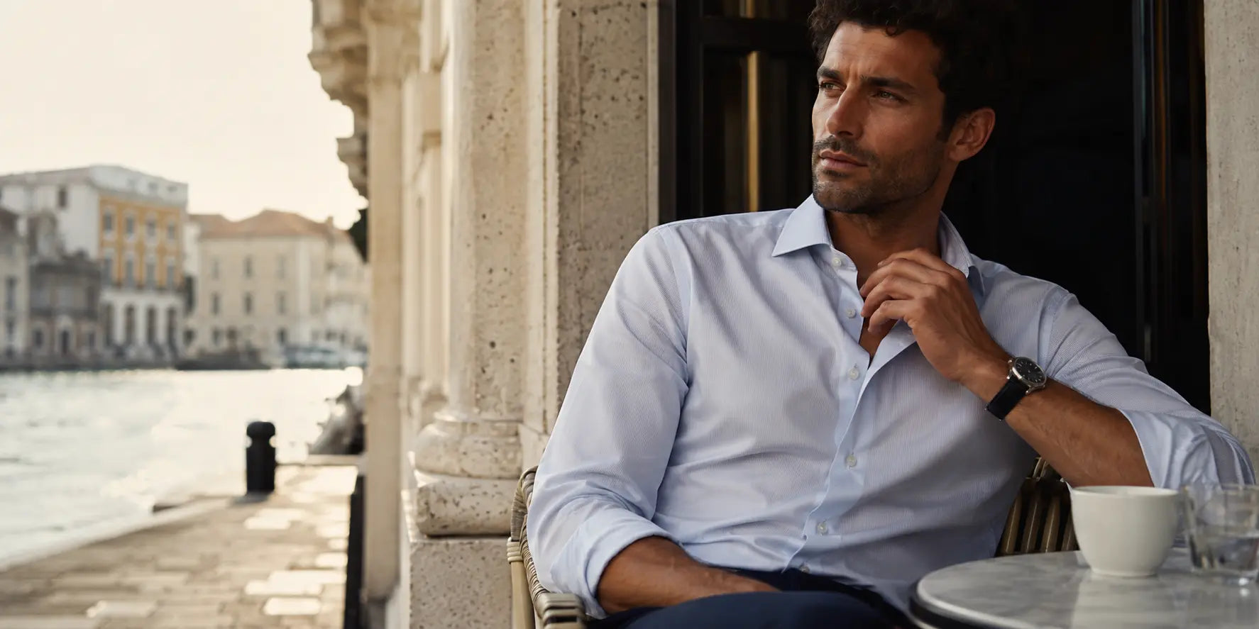 Hombre con camisa azul clara en una terraza elegante junto al agua, en imagen editorial para guía de camisas de hombre.