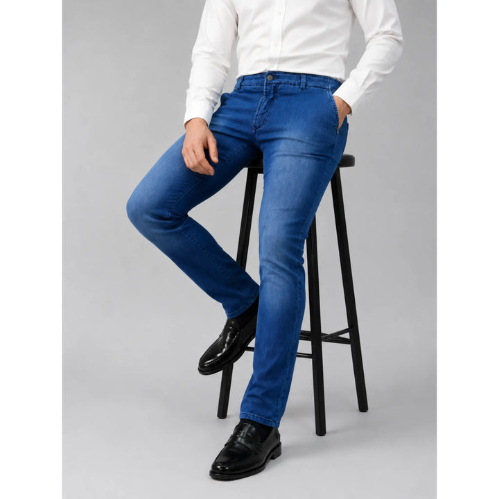 Pantalón vaquero tipo chino Santiago azul para hombre, sentado en taburete con camisa blanca y mocasines negros.